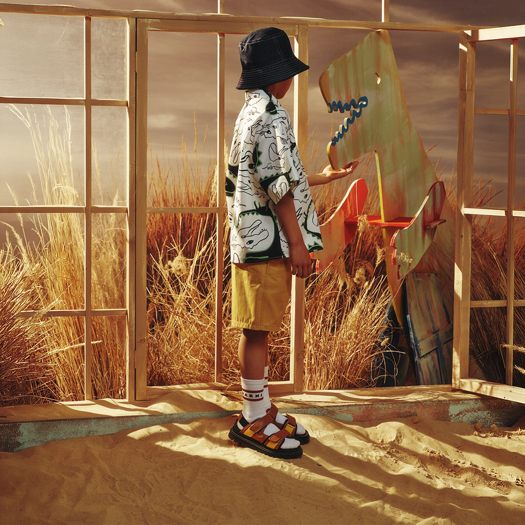 A child standing with a stylish outfit. The ground is full of sand and there is a window behind him with the sun that comes in