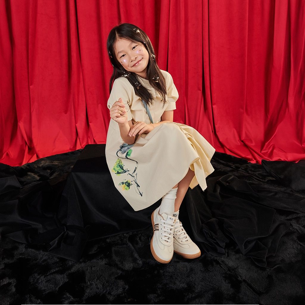 Young girl in a white dress with floral embroidery sitting on a black rug in front of red curtains.