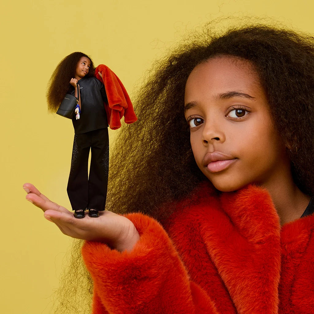 A girl with a miniature version of herself, wearing a red faux fur coat, against a bright yellow background.